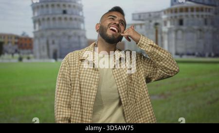 Giovane ispanico sorridente, mostra il gesto della mano accanto alla torre pendente dell'edificio di pisa in piazza all'aperto; viaggia divertente e divertente. Foto Stock