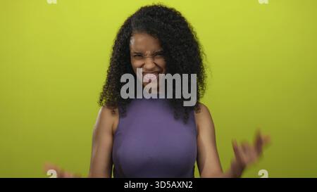 La giovane donna con i capelli ricci e le braccia alzate mostra il gesto delle corna di roccia in un ambiente giallo da studio; atmosfera divertente. Foto Stock