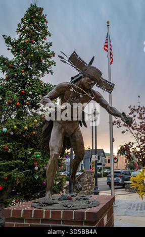 La scultura Nisenan Dancer di Douglas Van Howd nella Central Square di Auburn si erge accanto a un albero di Natale decorato alle prime luci delle festività serali. Foto Stock