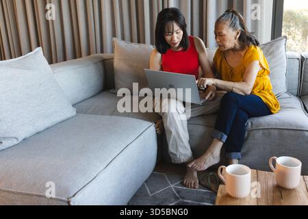 Sul divano, madre asiatica anziana e figlia che usano il laptop, discutono di qualcosa di importante Foto Stock