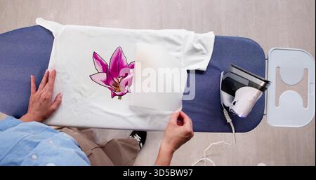 Donna che personalizza il design di una T-shirt con pressa per il trasferimento di ferro a casa. Foto Stock