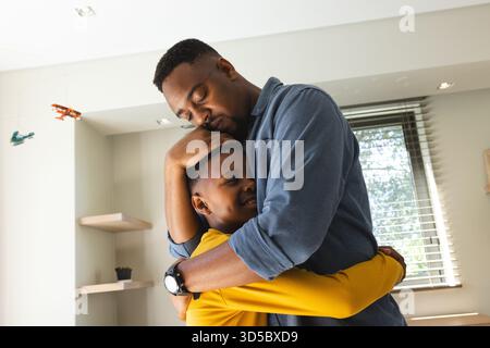 Padre e figlio afroamericani che condividono un caldo abbraccio in un accogliente soggiorno, godendosi momenti di sole Foto Stock