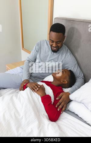 Padre e figlio afroamericani sdraiati sul letto nella camera da letto interna, sorridendo con una coperta a righe Foto Stock