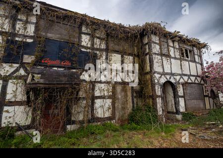 Edificio pub abbandonato con cornici in legno, aperture a bordo e una forte crescita eccessiva che si estende lungo la facciata in decomposizione Foto Stock