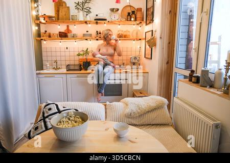 Una donna siede in una cucina con un bambino in braccio. La cucina è arredata con molta luce e ha un tavolo con una ciotola di popcorn e una tazza Foto Stock