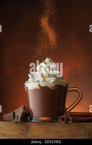 polvere cosparsa su una tazza di cioccolata calda con panna montata e cannella su sfondo marrone Foto Stock