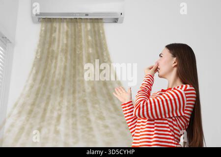 Giovane donna che si copre il naso a causa del cattivo odore del condizionatore d'aria al chiuso. L'aria sporca fluisce dall'apparecchio Foto Stock
