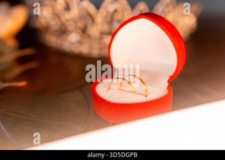 Un primo piano di due fedi nuziali in oro in una scatola di velluto a forma di cuore rosso, circondata da oggetti di gioielleria come una corona e un diamante su tessuti tradizionali. Foto Stock