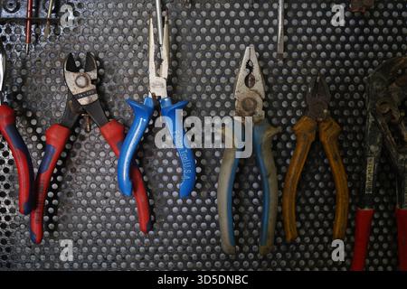 Utensili manuali assortiti, tra cui vari tipi di pinze e taglierine, appesi in modo ordinato su un pannello di metallo perforato nero in un'officina. Foto Stock