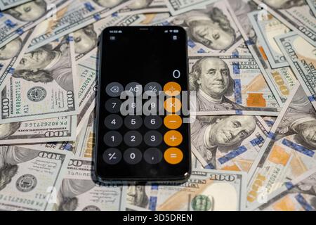 Uno smartphone mostra un'app di calcolo circondata da molte banconote da cento dollari, simboleggiando i calcoli finanziari. Foto Stock