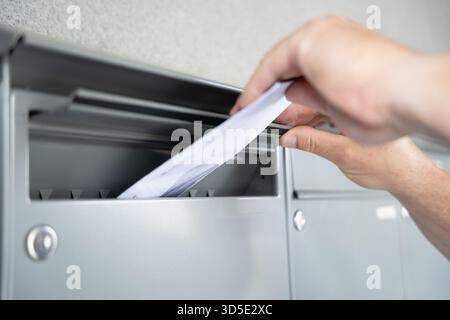 Primo piano di una mano umana che inserisce una busta bianca in una fila di cassette postali grigie in metallo Foto Stock