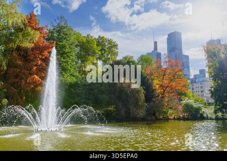 Francoforte, Germania - 21 ottobre 2025: Una vivace fontana spruzza acqua in un laghetto autunnale, incorniciato da alberi colorati. I moderni grattacieli di Francoforte Foto Stock