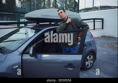 Un uomo si trova accanto a una piccola auto blu con un vano di carico sul tetto, regolando la giacca e preparandosi per un viaggio. La scena suggerisce viaggi, bagagli e jou Foto Stock
