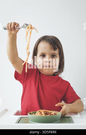 La bambina con la camicia rossa regge entusiasticamente la forchetta con gli spaghetti, seduto sul seggiolone con un piatto di pasta su sfondo bianco, godendosi Foto Stock