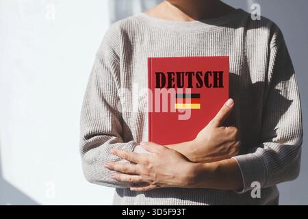 La giovane donna in maglione beige tiene un libro di testo in tedesco rosso con iscrizione tedesca e bandiera tedesca, concetto di apprendimento delle lingue straniere. la sua faccia io Foto Stock