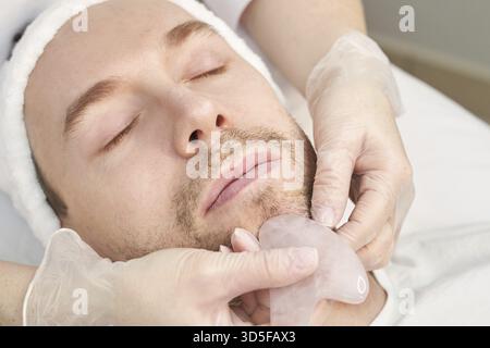 Medico che fa la routine cutanea. Trattamento di maternità in salone. Concetto di assistenza sanitaria. Uomo. Massaggio facciale pandemico con agopuntura guasha. Procedura anti-età Foto Stock
