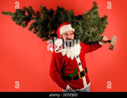 Babbo Natale con l'albero di Natale. Uomo barbuto nel cappello di Babbo Natale con l'albero di Natale. Bel maschio in costume di Babbo Natale che trasporta albero di Capodanno artificiale. Babbo Natale Foto Stock