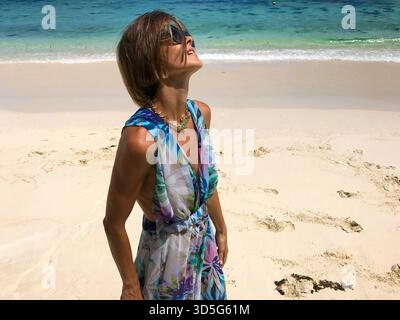 Foto a metà corpo di una bella giovane donna con un abito floreale estivo mentre si trova in piedi su idilliaca spiaggia tropicale con acqua blu turchese. Ritratto alla moda Foto Stock