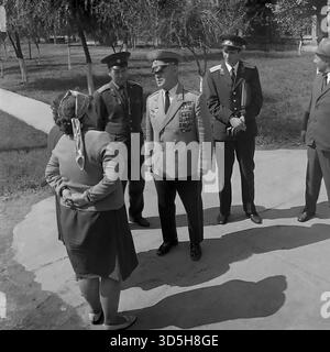 URSS, RSS Ucraina, Slavyansk, 1975. Un colpo sincero della celebrazione del 30° giorno della Vittoria, che si svolge in un vicolo della città vicino allo stabilimento "Krasny Metallist". Un veterano di guerra sovietico di alto rango e altamente decorato in uniforme da parata completa è il centro dell'attenzione. È in conversazione con i residenti locali, tra cui due donne (una in un velo). È accompagnato da altri due ufficiali militari e da un ufficiale civile con un cappello fedora. Questa foto archivistica in bianco e nero cattura gli ospiti onorati che interagiscono con i cittadini. Foto Stock