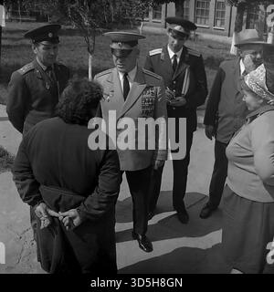 URSS, RSS Ucraina, Slavyansk, 1975. Un colpo sincero della celebrazione del 30° giorno della Vittoria, che si svolge in un vicolo della città vicino allo stabilimento "Krasny Metallist". Un veterano di guerra sovietico di alto rango e altamente decorato in uniforme da parata completa è il centro dell'attenzione. È in conversazione con i residenti locali, tra cui due donne (una in un velo). È accompagnato da altri due ufficiali militari e da un ufficiale civile con un cappello fedora. Questa foto archivistica in bianco e nero cattura gli ospiti onorati che interagiscono con i cittadini. Foto Stock