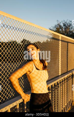 Atleta donna che si ferma dopo l'esercizio contro una recinzione durante il tramonto Foto Stock