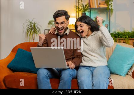Allegra giovane e variegata coppia in abiti casual che usa il computer portatile sorpreso con il jackpot di gioco online della lotteria che ha vinto per e-mail sul divano nel soggiorno di casa. La famiglia indiana felice stringe i pugni in appartamento Foto Stock
