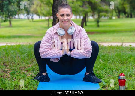 Donna sorridente che fa lo squat yoga posa sul tappeto all'aperto, indossa le cuffie, parco. Foto Stock