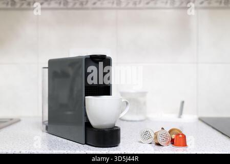 Moderna macchina da caffè a capsule da una tazza per preparare l'espresso in una tazza bianca su un bancone della cucina con cialde di caffè sparse. Foto di alta qualità Foto Stock