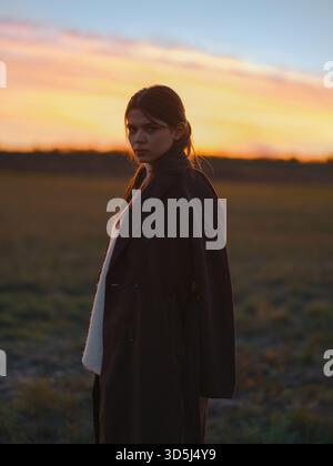 Giovane donna seria in piedi sul campo contro il suggestivo cielo arancione e rosa del tramonto. Indossa un cappotto scuro su un maglione bianco, guardando con fiducia il Came Foto Stock
