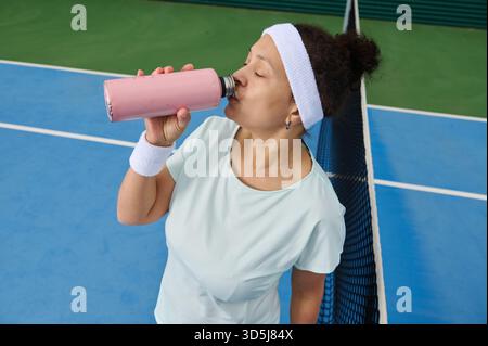 Una donna atletica si prende una pausa su un campo da tennis, sorseggiando un frullato rosa. Indossa un cerchietto bianco e un braccialetto, appoggiandosi casualmente alla rete W Foto Stock