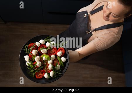 Una donna che indossa un grembiule tiene con cura un piatto nero ricoperto di insalata caprese fresca, con pomodori a fette, palline di mozzarella e basilico in un ambiente moderno da cucina. Foto Stock
