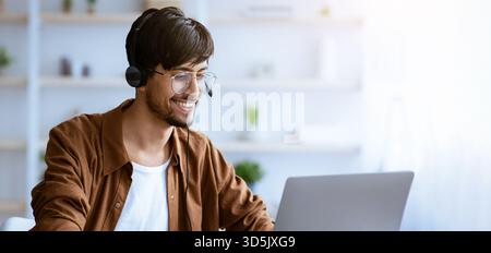 Giovane uomo che lavora a casa con notebook e cuffie durante la sessione di riunione online Foto Stock