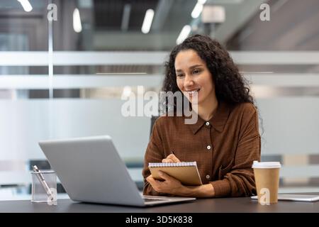 Donna d'affari con capelli ricci sorridenti e note scritte, in un notebook a spirale mentre partecipa a un webinar online o a una riunione su un notebook in un ufficio moderno Foto Stock