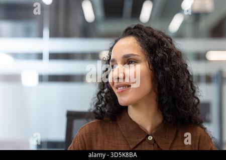 Giovane donna d'affari con capelli ricci sorridenti che esprimono speranza e positività, che guarda al lato con fiducia in un ambiente moderno open space Foto Stock
