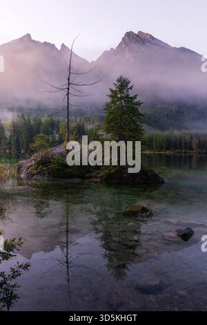 Montagne ricoperte di nebbia sopra un lago limpido con un'isola rocciosa e alberi che si riflettono nell'acqua ferma. Foto Stock