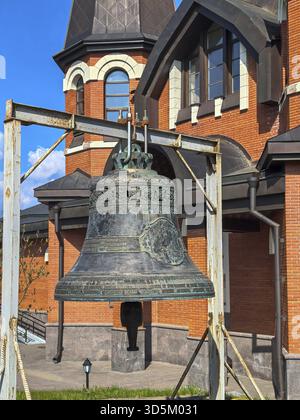 Yukki, Oblast' di Leningrado, Russia - 28 maggio 2025. Una grande campana dettagliata in primo piano, con la chiesa di San Giovanni Battista a Yukki Foto Stock