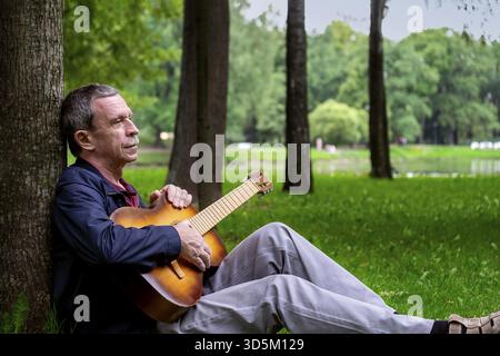In estate sera, l'uomo romantico adulto si siede nel bellissimo parco sull'erba, appoggiandosi contro l'albero, e suona la chitarra acustica classica a sei corde. Selettivo Foto Stock