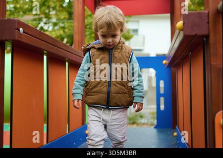 Un bambino in un gilet marrone cammina lungo un colorato percorso del parco giochi, catturando l'infanzia gioiosa, l'amore per la famiglia e la cura protettiva in un autunno luminoso Foto Stock