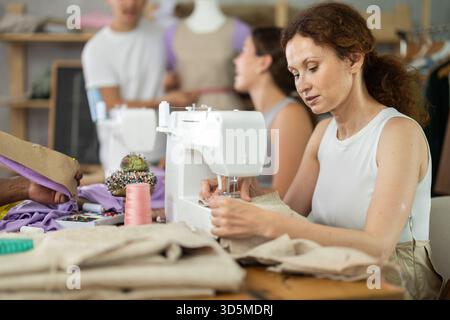 Donna di mezza età che cucisce mentre altre lavorano con macchina da cucire e manichino Foto Stock