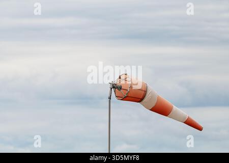 Giacca a vento orizzontale a righe bianche e arancioni su un palo metallico che soffia nel vento contro il cielo nuvoloso coperto. Ripresa da un angolo basso che mostra l'indicatore di direzione del vento Foto Stock