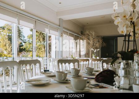 Elegante sistemazione interna per ricevimenti nuziali o eventi formali con sedie bianche, tavoli e grandi finestre da giardino Foto Stock