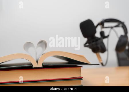 Un libro aperto con pagine piegate a forma di cuore si trova su libri impilati, con un microfono podcast sfocato sullo sfondo. Il simboleggia la creatività Foto Stock