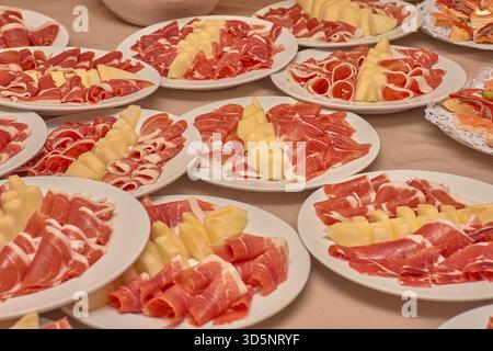 Dozzine di piatti bianchi contengono tartine di prosciutto stagionato accompagnate da fette di melone, disposte in file su un tavolo per un grande raduno. Foto Stock