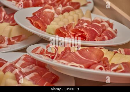 Dozzine di piatti bianchi contengono tartine di prosciutto stagionato accompagnate da fette di melone, disposte in file su un tavolo per un grande raduno. Foto Stock