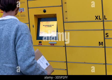 Person holt Paket an DHL-Packstation ab 16.11.25, Selters: Symbolfoto, Illustrationsbild, Symbolbild, Illustrationsfoto, Alltagsszene Eine person steht mit einem kleinen Paket vor einer gelben DHL-Packstation und blickt auf den Bildschirm, der den Begrüßungsbildschirm Willkommen bei der Packstation anzeigt. Die Szene wirkt alltäglich und zeigt die Nutzung eines automatisierten Paketautomaten im öffentlichen Raum. Der Fokus liegt auf dem Bedienfeld der Packstation und dem Paket in der Hand der Person. Selters im Taunus Hessen Deutschland *** la persona ritira il pacco alla stazione DHL Packstation 16 11 25, S. Foto Stock