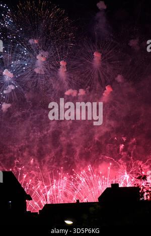 I vivaci fuochi d'artificio si esibiscono nel cielo notturno, con raffiche rosa e viola con fumo e sagome dei tetti in primo piano Foto Stock