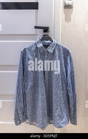 Una camicia blu a maniche lunghe da uomo appesa su un appendiabiti su un pomello della porta su una porta bianca Foto Stock