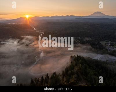 Vista aerea del sole che sorge all'orizzonte e di un fiume con nebbia che scorre attraverso la verde valle di Orting, Washington, Stati Uniti. Foto Stock