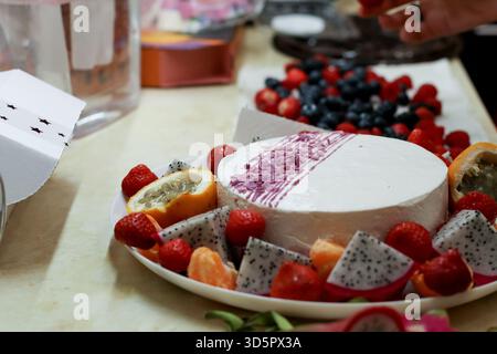 Vivace assortimento di frutta a fette e frutti di bosco disposti intorno a un dessert bianco rotondo con salsa viola su un piatto in cucina Foto Stock