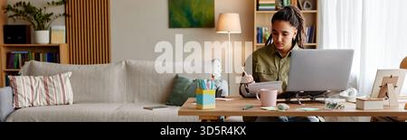 Giovane donna adulta che studia a casa utilizzando un notebook e prendendo appunti Foto Stock